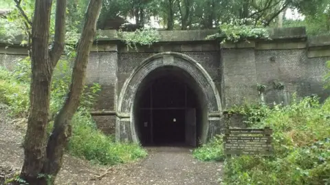 Malc McDonald/Geograph Kelmarsh Tunnel, showing gates to a brick structure tunnel, with an archway. There are trees and bushes around the entrance and a path leading into the tunnel. A wooden sign, with writing on, is to the right. Trees and foliage are above the tunnel. 