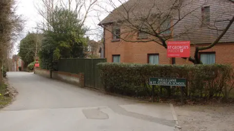 Alan Hunt/Creative Commons A picture of the entrance leading to a school. There is a two-storey brick building with wooden slats on one section to the right of the entrance behind a hedge and a red sign that reads ' St George's Ascot '. In front of the hedge is a green and white street sign with the words Wells Lane & St George's School.