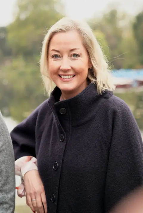 BBC/Graham Hodson Survivor Gemma, a blonde woman, pictured standing in a park and smiling at the camera. She's wearing a black winter cape with a blush blouse. 