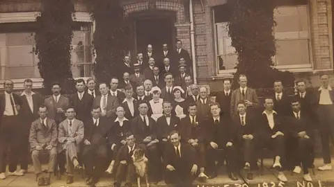 Kitchener's Guest House A black and white image of the mostly male staff at Kitchener's Memorial Home in Lowestoft, dated 30 June 1925. There are roughly two rows of people, the first row seated and the second row standing, with two women wearing servants' white caps and aprons, in the middle. There are more men standing in a group behind, gathered on stairs up of the front door or a Victorian building. The men are wearing suits, some with ties and others without. Sitting on the ground are two more men and a dog. 