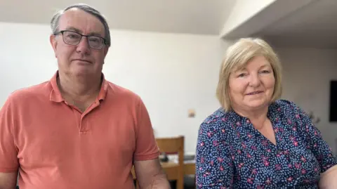 A man in late middle age with short grey hair and wearing a coral-coloured polo shirt and glasses sits at a kitchen island next to a woman of a similar age, with a short blonde bob and wearing a blue dress with small pink and light blue flower pattern. A dining table and a chair are just visible in the background and the walls are painted cream. 