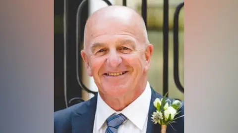 A photo of Barry Howard. He is facing the camera and smiling. He is bald and is wearing a blue suit with a blue tie. 