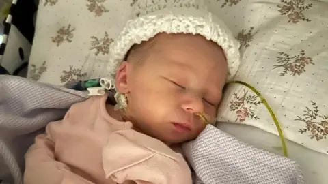 Tom A sleeping baby wearing a white had and pink top.