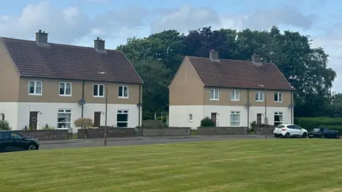 Some of the houses in Linburn Estate - a few of which are now lying empty. They are painted cream on the bottom half, brown on the first floor. Each has a brick garden wall and the grass in front is well-kept.