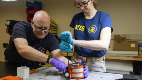 NTSB Male in Lickent Gloves Nurswe Gloves carrying the black box orange casing, while employee uses the drop tool to open its casing.