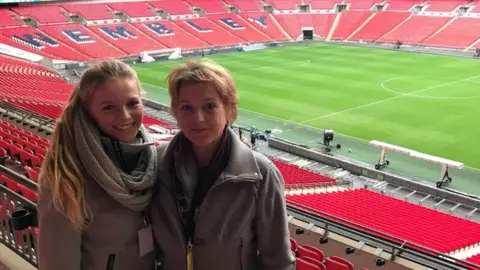 Nadine Noordam A woman and her daughter smile at the camera as they stand for a photo in the stands at Wembley, with the pitch in the background behind them