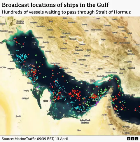 A satellite image of the Strait of Hormuz with ships marked with arrows. It shows hundreds of ships clustered by the strait waiting to pass. It is timestamped to 09:39 BST on 13 April