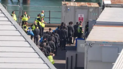 PA Media A group of migrants with backs to camera in a queue to board a bus in the Border Force compound in Dover, Kent