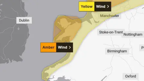 A UK weather warning map showing wind alerts along the west of the country. A wide yellow-shaded area marked “Yellow – Wind” stretches from southwest Wales up the Welsh coastline and into northwest England, reaching toward the Manchester area. Within this zone, a smaller, darker orange-shaded region labeled “Amber – Wind” covers parts of western Wales near the coast, indicating a higher level of warning. 
