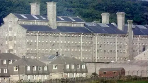 The five-story high grey granite main building of Dartmoor prison, with houses in the foreground and moorland in the background.