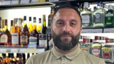 A man with dark hair and a beard standing in front of shelves full of alcohol