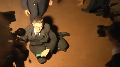 A man in a dark coloured suit sits on the floor among a group of people. There is a bright light focused on his face and a uniformed police officer stands behind him. 