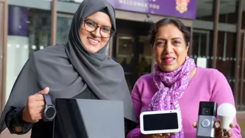 City of Wolverhampton Council Obaida and Jaspal are holding several devices that are being deployed in the pilot scheme. They are standing outside a building. Obaida has glasses and a grey hijab. Jasbir is wearing a pink top and cardigan.