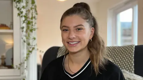 Beth sits in a cushioned armchair in a living room, wearing a black sports-style top. Shelving with plants and decorative items is visible behind them, along with soft indoor lighting.