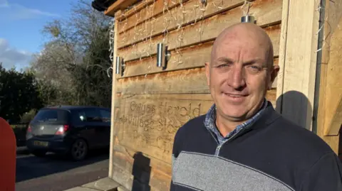 BBC A bald man wearing a navy blue fleece is pictured outside a wooden cabin during a sunny day. 