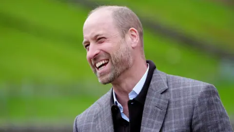Owen Humphreys/PA The Prince of Wales wearing a grey suit jacket, black swearer and white shirt. He is laughing at something off camera. The background is blurred but seems to be green grass and hills.