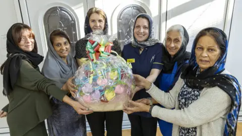 Telford and Wrekin Council Six women, all wearing head scarves, have their arms outstretched in front of them and are holding a basket filled with colourful knitted hearts. The basket is wrapped in plastic and tied with a colourful ribbon