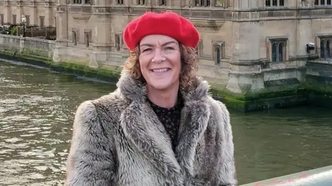 Sharon Doherty is wearing a red beret and fur coat.
She has short curly hair and is standing beside a river with a historic-looking building in the background.