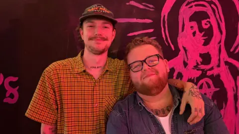 Two men pose together in front of a black wall featuring pink artwork. On the left, a man wears a yellow checked shirt and cap; on the right, a man wears glasses and a denim shirt.