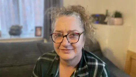Sarah is sitting on a grey ribbed sofa and is wearing a green, black and grey checker shirt and wearing red black and brown turtle shell glasses. The background is blurred. 