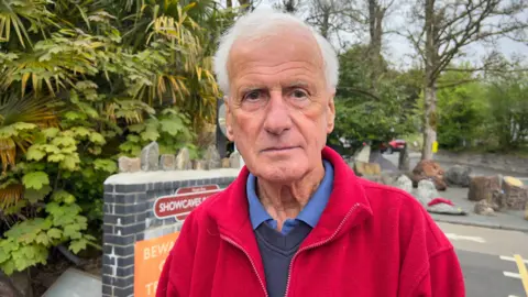 Ashford Price, Chairman of the National Showcase Centre at Dan yr Ogof Caves, stands in front of the entrance looking at the camera. He is in a red jacket with the trees and ferns surrounding the entrance behind him
