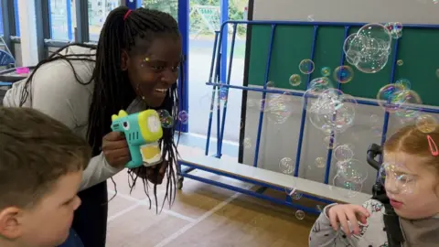 A woman using a bubble machine to amuse two children. They are in an indoor sports hall and there is a large window behind them.