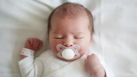 A newborn baby lying in a cot with a dummy in their mouth. They are wrapped in white and still have a hospital tag wrapped around their hand.