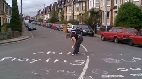 Playing Out A woman chalks the words "children playing" on a suburban street.