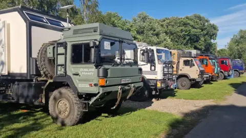 The Vanlife Festival Seven large trucks are parked on a patch of grass with trailers on the back of the trucks.