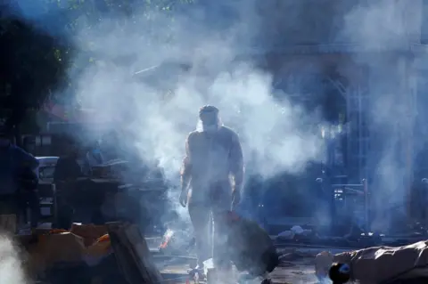 Adnan Abidi / Reuters A family member wearing personal protective equipment (PPE) stands next to a body of a person in crematorium as smoke hangs in the air