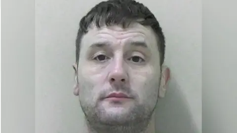A police mugshot of Gary Rarity, a man in his 40s with stubble, dark eyes, and a dark fringe-style haircut with shaved back and sides.
