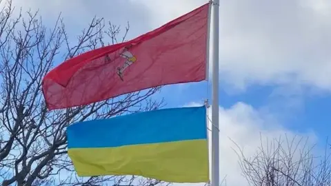 A flagpole with the Manx flag and the Ukrainian flag beneath blowing in the wind.