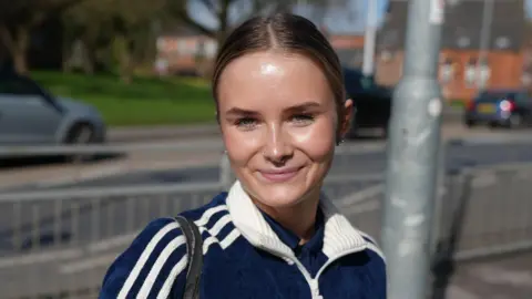 A woman with brown hair tied up. She is smiling and has blue eyes. She is wearing a blue tracksuit top with with stripes down the arms and a white collar. Cars can be seen passing on a road in the background.