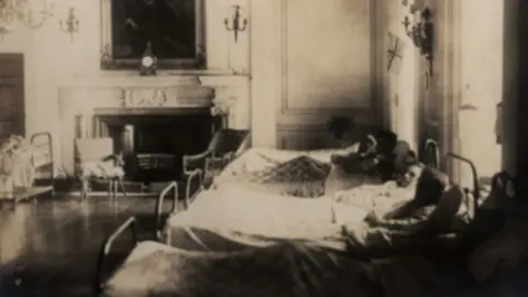 North Lincolnshire Museums A grainy, black and white image showing a former ballroom with a few hospital beds in it. Four of the beds contain men. A chandelier hangs near the beds.