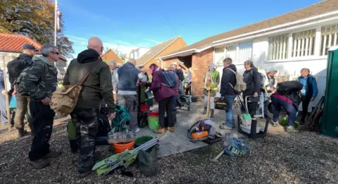 Volunteers who are part of the dig are huddling around the village hall collecting equipment. Orange buckets, chairs and spades are strewn on the floor as people begin to collect their things.