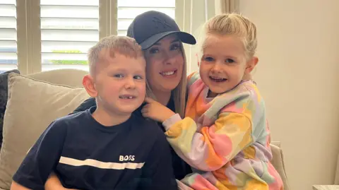 Ashley, who is wearing a black baseball cap and black T-shirt, is sitting on the couch in her home in front of a window with her two children. They are all smiling at the camera.