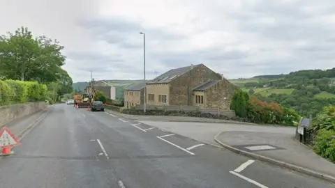 A T-junction on a rural road. Properties with sloped rooves can be seen in the foreground at the junction, with rolling hills in the far distance. Parked vehicles are further down the road.