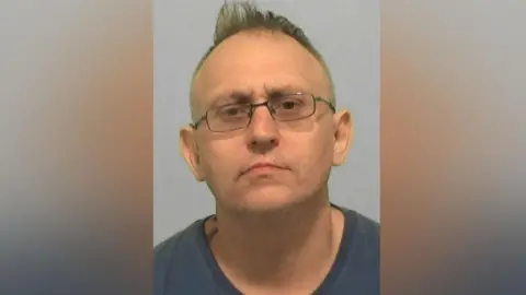 Northumbria Police Mugshot of Colin Williams. He is clean shaven, is wearing thing glasses and has a quiff of blond hair sticking up atop his balding head