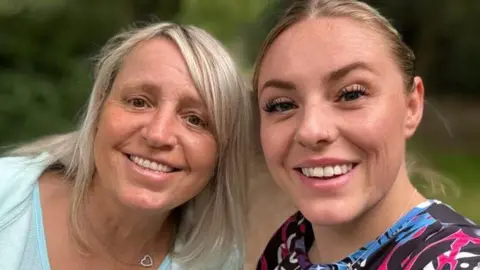 Georgina King Two women smile at the camera as the one on the right takes a selfie of them. They both have long blonde hair - the one on the left has a blue top, the one on the right has a patterned black, purple and blue top,