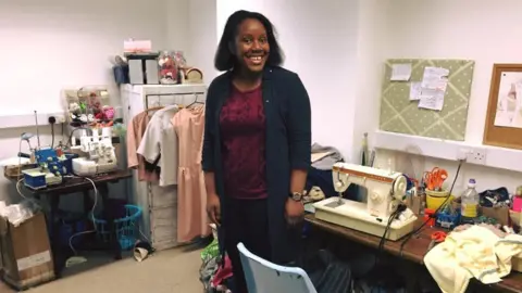 Emergence Restock Shalize is standing in a work room with two tables behind her with sewing machines and items related to sewing such as scissors and a heap of yellow material. She is smiling to camera and is wearing a burgundy top with a long black cardigan.