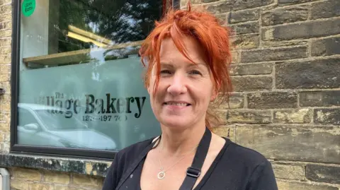 A woman wearing a black top and apron standing outside a sandwich shop.