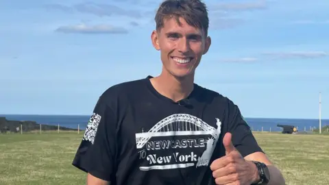 Leodhais Macpherson smiling at the camera and making a thumbs-up gesture after arriving in Tynemouth. He has short brown hair and is wearing a black t-shirt with a picture of the Tyne Bridge and the words Newcastle to New York 2025. He is standing on a grassy area with the sea behind him.