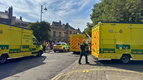 Several yellow‑green NHS ambulances and an incident response unit are parked on a street, with an ambulance worker in uniform at the rear of one vehicle and people standing nearby beside historic stone buildings under a clear sky.