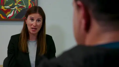BBC Reporter Helen Drew wearing a dark blazer speaks during an interview, filmed over the shoulder of the person opposite her