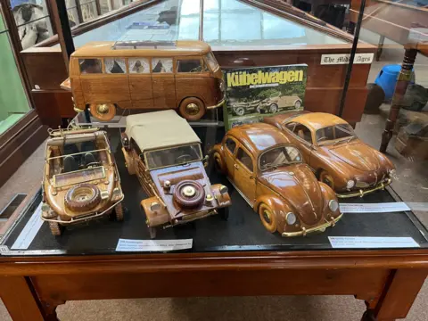 John Devine/BBC A collection of four Volkswagen vehicles sit in a display case in the main gallery of a museum, including the iconic camper van and Beetle, models are highly polished wood and brass about 30cm long and 15cm high they are very detailed.