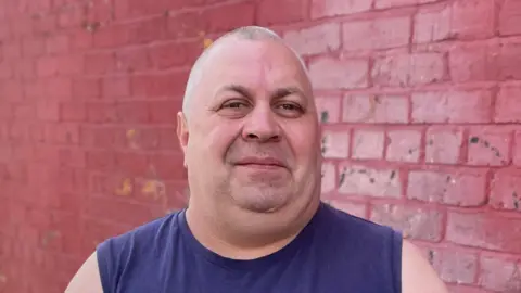 Mark Ansell/BBC A bald man with a blue vest smiles at the camera. He is standing against a background of red bricks