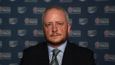 John Fairhall/BBC A man with short greay hair and a trimmed beard wearing a suit and tie looking at the camera. Behind him is a blue branded banner or backdrop with the Essex Police crest and lettering on it. 