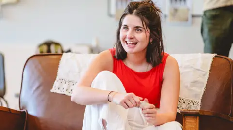 Andrew Billington Isabella has a red top on and white trousers. She has medium length brown hair. She is sat in a big, brown chair smiling at the camera.