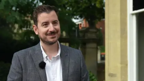 Mike Reader has brownish hairsbreadth and a beard wearing a grey suit overgarment and achromatic shirt. He has a microphone pinned to his jacket. He is opinionated extracurricular successful beforehand of trees and to nan correct of him is nan flimsy position of nan exterior of a beige building.