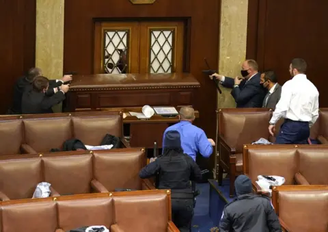 Drew Angerer / Getty Images Capitol police officers point their guns at a door that was vandalized in the House Chamber during a joint session of Congress on 6 January 2021 in Washington, DC.